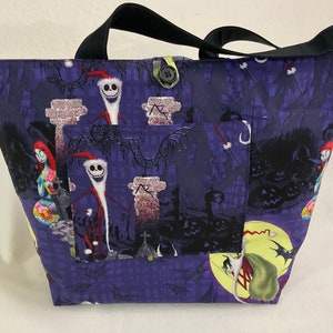 May include: A tote bag featuring a dark purple fabric with a spooky Halloween design. The bag has a black handle and a pocket on the front. The design includes characters from The Nightmare Before Christmas.