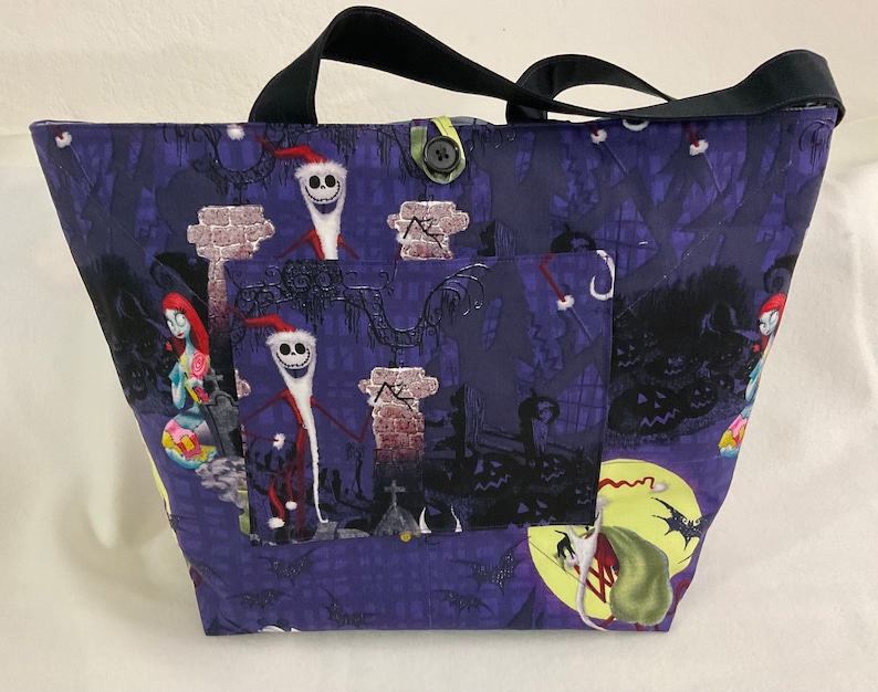 May include: A tote bag featuring a dark purple fabric with a spooky Halloween design. The bag has a black handle and a pocket on the front. The design includes characters from The Nightmare Before Christmas.