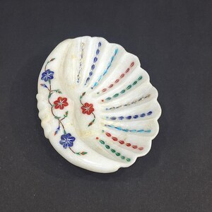 Peut inclure: Plat en forme de coquillage en marbre blanc avec des motifs floraux et linéaires incrustés. Le plat présente des accents bleus, rouges et verts, avec une vigne florale sur le côté gauche. Bord festonné. Décoratif et fonctionnel.