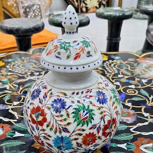 May include: A white ceramic jar with a lid decorated with intricate floral patterns made from inlaid colorful stones. The jar is sitting on a black and white marble surface.