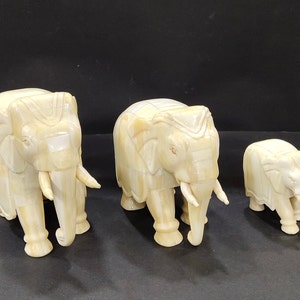 Op de afbeelding: Drie ivoorkleurige olifantenbeeldjes van verschillende groottes. De olifanten hebben gedetailleerde gravures en zichtbare slagtanden. De grootste olifant is ongeveer 20 cm hoog, de middelste 15 cm en de kleinste 10 cm.
