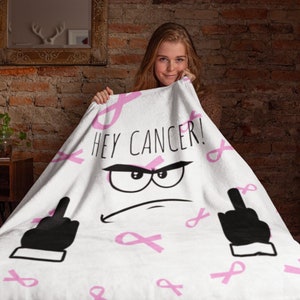 May include: White blanket with pink ribbon accents and the text "HEY CANCER!" in black. The blanket features a cartoon face with an angry expression and two black middle fingers. The blanket is a statement piece.