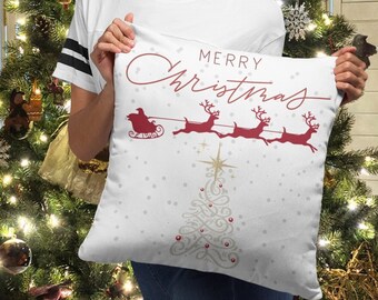 Puede incluir: Almohada blanca con un diseño rojo de "Feliz Navidad" que presenta una silueta de Papá Noel en un trineo tirado por renos. La almohada está decorada con una estrella dorada y un pequeño árbol de Navidad.