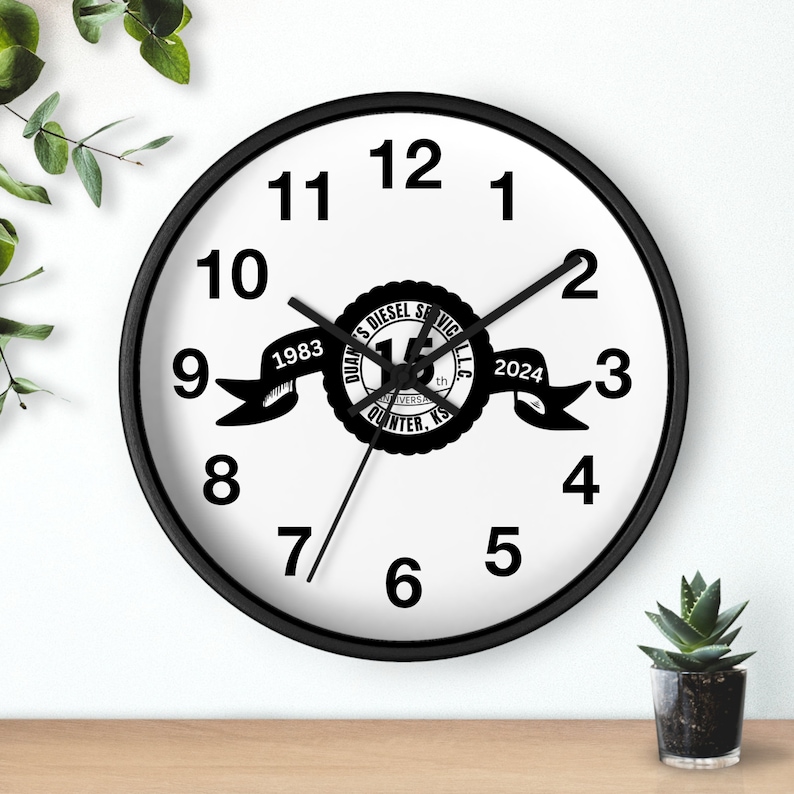 May include: Black and white wall clock with black hands. The clock face has black numbers and a black and white logo that says "Quan's Diesel Service LLC 15th Anniversary 1983-2024 Quinter, KS".