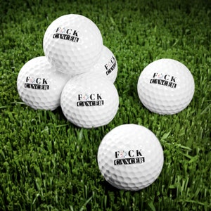May include: Five white golf balls with the phrase "F*CK CANCER" printed in black on each ball. The golf balls are on a green grassy surface.
