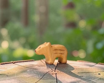 May include: A small, light brown wooden figurine carved in the shape of a wild boar piglet. The figure features a smooth, rounded body with carved horizontal stripes along its side and a simple, defined snout, standing on four short legs.