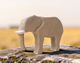 May include: A light brown wooden elephant figurine with a smooth, matte finish. The animal figure features a single white painted tusk and white painted details on the feet, showcasing a simplified, carved silhouette with a downward-curving trunk.