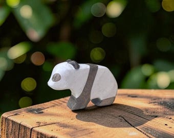May include: A small, hand-carved wooden panda figurine featuring a white body with painted dark gray patches around the eyes, ears, and across the midsection. The panda is depicted in a standing position with a smooth, matte finish on the wood.