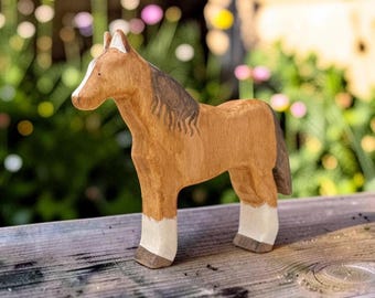 May include: A hand-carved wooden horse figurine featuring a light brown body, a darker brown mane and tail, and white markings on the lower legs. The horse has a white blaze on its face and dark brown hooves, showcasing a smooth, natural wood grain finish.