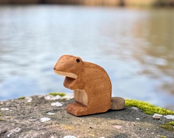 May include: A small, hand-carved wooden figurine of a beaver in a seated position. The figure features a smooth, medium-brown finish on the body and head, with a lighter, natural wood-toned chest and a flat, rounded tail extending from the back.