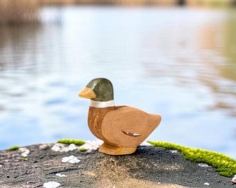May include: A small, hand-carved wooden duck figurine featuring a smooth, natural wood-toned body, a painted dark green head, a white neck ring, and a light yellow beak. The duck is depicted in a standing position with a simple, rounded shape.