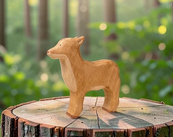May include: A light brown wooden figurine of a young deer standing in profile. The animal figure features smooth, rounded edges, a simple carved head with small ears, and a visible wood grain texture throughout the natural light brown surface.