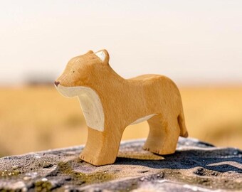 May include: A carved wooden figurine of a lioness in a standing position. The figure features a natural light brown wood grain finish with a smooth, rounded silhouette, a painted white chest area, and subtle dark brown details for the nose and eyes.