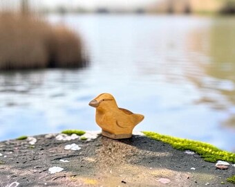 May include: A small, light brown wooden figurine shaped like a duck. The carving features a smooth, rounded body, a defined beak, and a simple wing detail on the side. The wood grain is visible throughout the surface of the duck-shaped object.