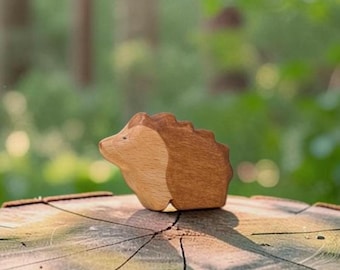 May include: A small, stylized wooden hedgehog figurine crafted from two distinct tones of wood. The front section is a light, natural beige wood, while the back section is a darker, rich brown wood with a serrated, textured edge representing quills.