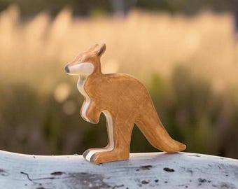 May include: A wooden kangaroo figurine featuring a smooth, light brown finish with a cream-colored chest and face. The animal is depicted in a standing profile position with its tail extended backward and its front paws tucked near its chest.