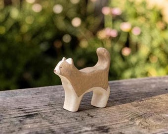 May include: A small, hand-carved wooden cat figurine featuring a two-tone design with a light beige upper body and a natural cream-colored lower body. The cat is depicted in a standing position with its tail curved upward and a simple painted face.