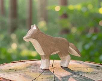May include: A stylized wooden figurine of a fox standing in profile. The fox features a smooth, light brown coat on its back and head, with a contrasting cream-colored chest, underbelly, and lower legs. The ears are pointed and the tail is bushy.