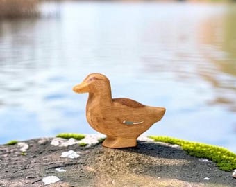 May include: A small, hand-carved wooden duck figurine with a smooth, light brown finish. The duck features a simple, stylized shape with a rounded head, a flat beak, and a small, painted blue and white detail on the side of its wing area.