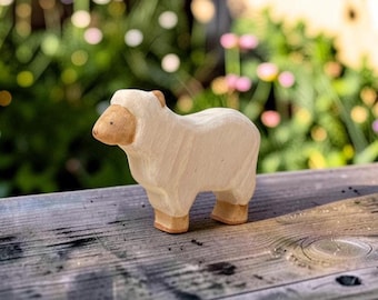 May include: A hand-carved wooden sheep figurine featuring a light cream-colored body with a smooth finish. The sheep has a darker tan-colored face and matching tan-colored hooves, with a simple, minimalist shape and visible natural wood grain.