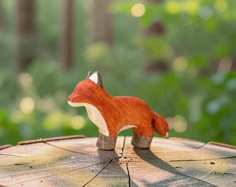 May include: A small wooden fox figurine featuring a vibrant orange coat, a white chest and muzzle, and dark brown paws. The fox is depicted in a standing position with its head turned slightly to the side, showcasing smooth, hand-carved details.