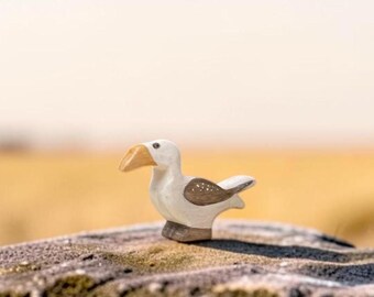 May include: A small wooden bird figurine featuring a white body, a light brown beak, and brown wings with white dot details. The bird is depicted in a standing position with a smooth, carved finish and a dark brown base.