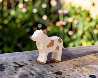 May include: A small, hand-carved wooden figurine of a calf. The animal is painted in a creamy off-white color with distinct light brown patches on its side and back. It features a brown ear, a brown snout, and brown hooves on all four legs.