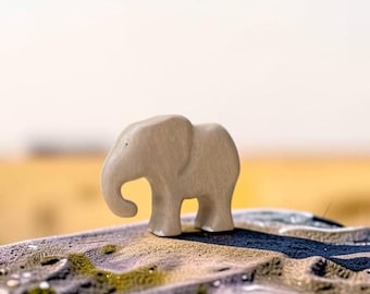 May include: A small, light beige wooden elephant figurine with a smooth, matte finish. The elephant is depicted in a standing position with its trunk curved downward, showcasing a simple, stylized silhouette carved from a single piece of wood.