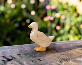May include: A small, hand-carved wooden duck figurine with a smooth, light beige body and a distinct yellow-orange beak and webbed feet. The duck is depicted in a standing position, showcasing the natural wood grain texture throughout its form.