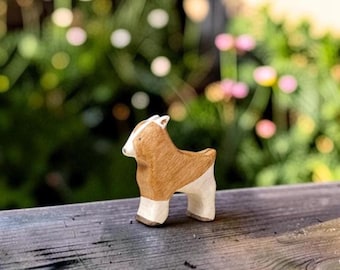 May include: A small wooden animal figurine shaped like a goat, featuring a light brown body with a white face, white lower legs, and a dark brown nose. The figure has a smooth, carved texture and stands on four legs, displaying a natural wood grain finish.