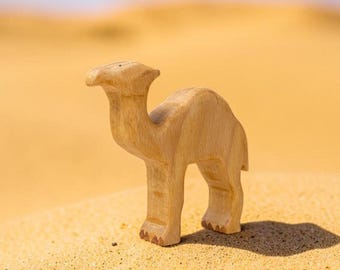 May include: A light brown wooden camel figurine with a smooth, carved surface. The animal is depicted in a standing position with a single hump on its back, a long neck, and a small head. The wood grain is visible throughout the entire structure.