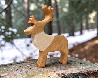 May include: A hand-carved wooden figurine of a reindeer standing in profile. The animal features a light tan head and antlers, a cream-colored chest, and a medium brown body. The wood grain is visible throughout the smooth, polished surface.