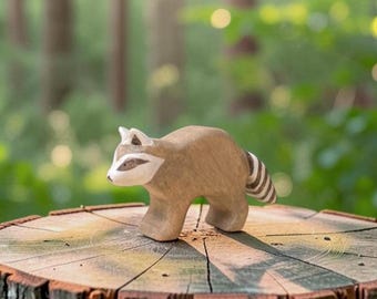 May include: A small, hand-carved wooden raccoon figurine featuring a smooth, light brown body, a white face with dark brown eye patches, and a striped tail. The animal is depicted in a standing position with its head turned slightly to the side.