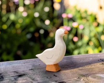 May include: A small, hand-carved wooden figurine of a goose. The body is painted white with a simple curved line indicating a wing, while the beak and the base are painted in a solid, earthy orange-brown tone. The figure has a smooth, matte finish.