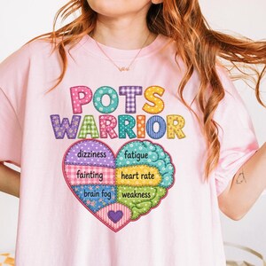 Pode incluir: Uma t-shirt rosa claro com o texto POTS WARRIOR em letras coloridas com padrões sobre um gráfico em forma de coração. O coração está dividido em secções com as palavras dizziness, fainting, brain fog, fatigue, heart rate e weakness.
