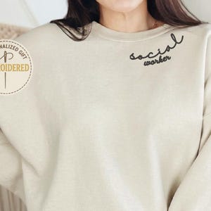 Pode incluir: Camisola bege com as palavras "social worker" em escrita preta. A camisola tem mangas compridas e gola redonda. O texto "PERSONALIZED GIFT EMBROIDERED" está impresso num autocolante circular no lado esquerdo.