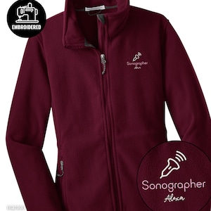 Puede incluir: Chaqueta de forro polar color granate con cremallera completa y cuello alto. La chaqueta presenta el texto bordado "Sonographer Alexa" y un gráfico de una sonda de ultrasonido. Un parche circular con la palabra "EMBROIDERED" es visible.