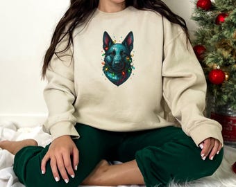 Op de afbeelding: Beige sweatshirt met een afbeelding van een Duitse herdershond versierd met kerstverlichting. De kop van de hond is turquoise en zwart, met rode en groene lampjes. De persoon draagt een groene joggingbroek.