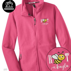 Può includere: Giacca in pile rosa con cerniera intera e due tasche laterali. La giacca presenta un ricamo a forma di ape con il nome "kayla" sotto. È presente anche la parola "ricamato".