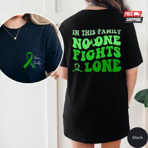 Może przedstawiać: Czarny T-shirt z zielonym tekstem, który brzmi "W tej rodzinie nikt nie walczy sam". Z tyłu koszulki znajduje się zielona wstążka z tekstem "Team Emily".