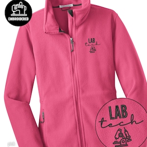 May include: A pink fleece jacket with a full zipper and two side pockets. The jacket has the embroidered text "LAB tech" with a microscope and syringe design. The upper left corner has a black circle with the word "EMBROIDERED".
