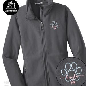 Puede incluir: Chaqueta de forro polar gris hierro con cremallera completa y cuello alto. La chaqueta presenta un diseño bordado de huella de pata con el nombre "Kayla" y "DVM" en escritura rosa. La chaqueta también tiene dos bolsillos laterales.