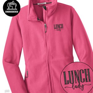 May include: A cerise fleece jacket with a full zip and two side pockets. The jacket features the embroidered text "LUNCH lady" on the chest. A circular patch with the same text is visible on the lower right. The jacket also has the word "EMBROIDERED" and a sewing machine icon.