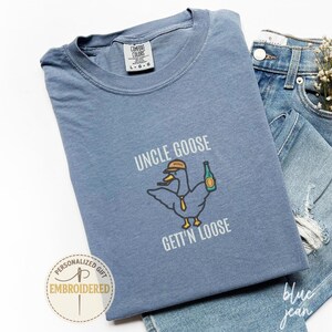 以下が含まれることがあります： 「UNCLE GOOSE GETT'N LOOSE」の文字と、帽子とボトルを持ったガチョウの漫画が描かれた、青色のComfort Colors Tシャツ。青いジーンズと組み合わせています。「PERSONALIZED GIFT EMBROIDERED」のステッカーも表示されています。
