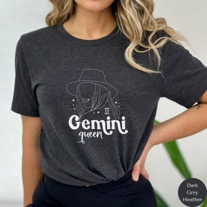May include: Dark grey heather t-shirt with a white graphic of a woman wearing a hat and the text "Gemini queen" with the Gemini astrological symbol.