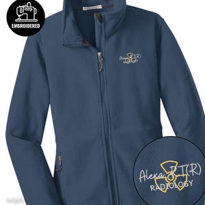 May include: Insignia blue fleece jacket with a full zipper and a high collar. The jacket has the embroidered text "Alexa RT(R) RADIOLOGY" in white and yellow. A black circle with the word "EMBROIDERED" and a sewing machine icon is in the upper left corner.