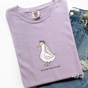 May include: A lilac-colored t-shirt with a cartoon duck wearing a floral crown and veil. The text "Wifey Honk Mode: Activated" is below the duck. The shirt is folded on top of blue denim jeans.