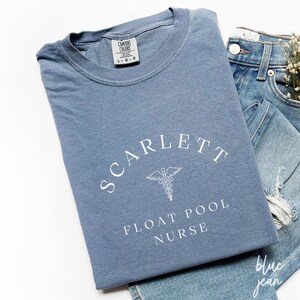 Pode incluir: Uma camiseta azul com texto branco que diz "SCARLETT FLOAT POOL NURSE" e um símbolo médico branco.