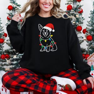 Peut inclure: Sweat-shirt noir avec un dessin blanc d'un Corgi portant un bonnet de Père Noël et des guirlandes lumineuses. La personne porte un pantalon de pyjama à carreaux rouges et noirs et des chaussettes blanches avec un motif de renne. Décorations de sapin de Noël en arrière-plan.