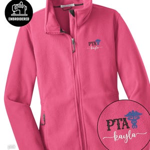 May include: A pink fleece jacket with a full zipper and two side pockets. The jacket has an embroidered design on the chest that says "PTA kayla" with a medical symbol. The upper left corner has a circular logo that says "EMBROIDERED".